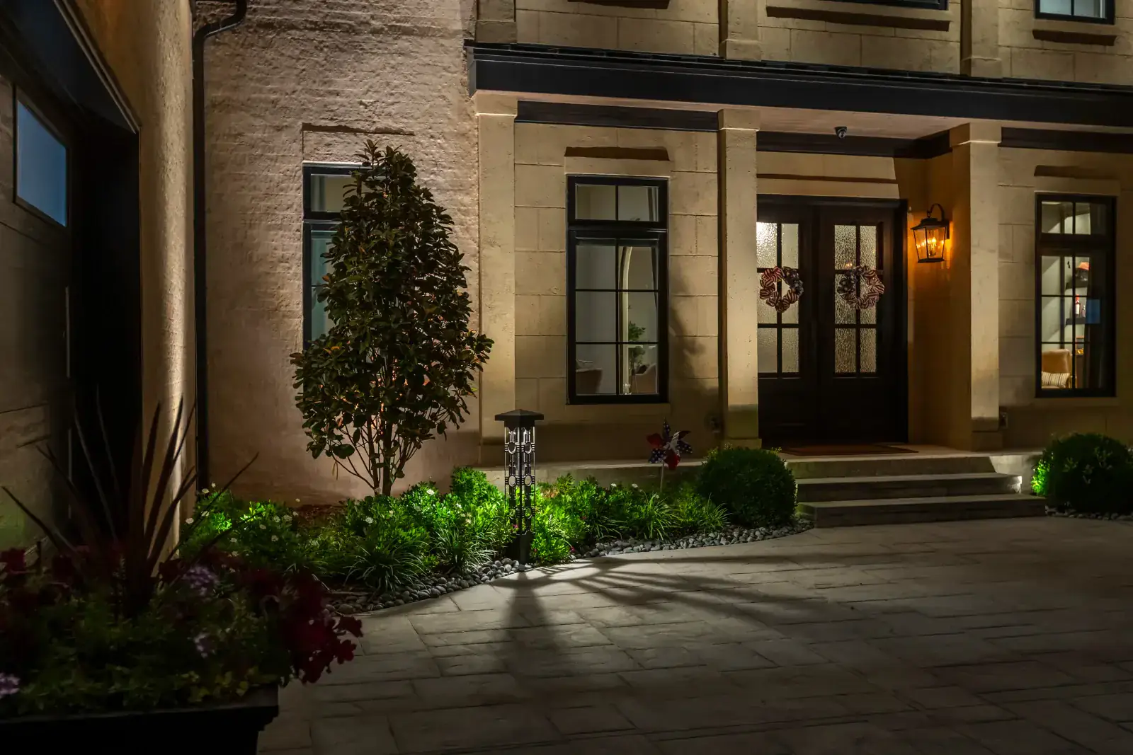 Architectural lighting illuminating a limestone home entry with uplighting on columns, garden plantings, and a paver driveway at night in Middle Tennessee by Light TN
